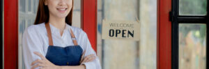 Asian woman in an apron stands holding a restaurant's menu in front of the door with a sign opening a shop, an opening of a small restaurant, a woman running a business. Operating a small restaurant.