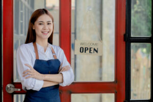 Asian woman in an apron stands holding a restaurant's menu in front of the door with a sign opening a shop, an opening of a small restaurant, a woman running a business. Operating a small restaurant.