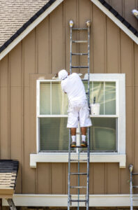 Painter man in white clothes with a protective scarf on his head paints a wooden window trim with a brush while standing on a high ladder leaning against the facade of a residential building