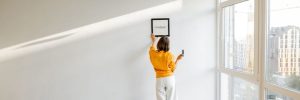 young woman hanging picture frame in room, decorating her newly renovated apartment, stands with her dog in white room
