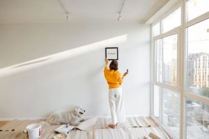 young woman hanging picture frame in room, decorating her newly renovated apartment, stands with her dog in white room